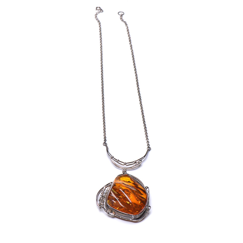 A close-up image of a genuine amber pendant showcasing its warm, golden hues. The amber is transparent with natural inclusions, capturing its organic beauty. The pendant is encased in a silver or gold frame, adding elegance to the raw, fossilized resin. The smooth surface of the amber reflects light, highlighting its natural glow and intricate details. This timeless piece of jewelry hangs delicately from a chain, blending nature and craftsmanship.