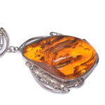 A close-up image of a genuine amber pendant showcasing its warm, golden hues. The amber is transparent with natural inclusions, capturing its organic beauty. The pendant is encased in a silver or gold frame, adding elegance to the raw, fossilized resin. The smooth surface of the amber reflects light, highlighting its natural glow and intricate details. This timeless piece of jewelry hangs delicately from a chain, blending nature and craftsmanship.