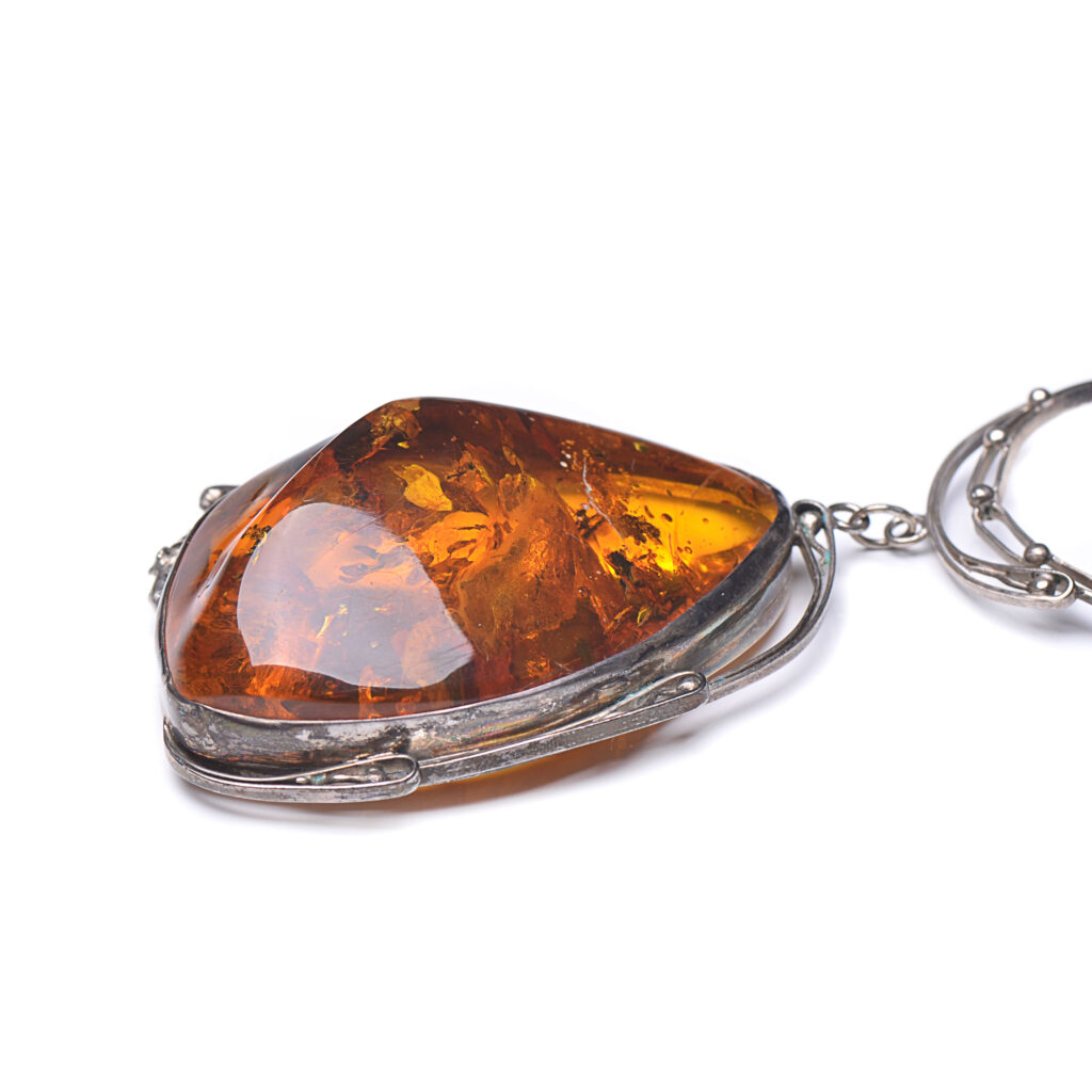 A close-up image of a genuine amber pendant showcasing its warm, golden hues. The amber is transparent with natural inclusions, capturing its organic beauty. The pendant is encased in a silver or gold frame, adding elegance to the raw, fossilized resin. The smooth surface of the amber reflects light, highlighting its natural glow and intricate details. This timeless piece of jewelry hangs delicately from a chain, blending nature and craftsmanship.