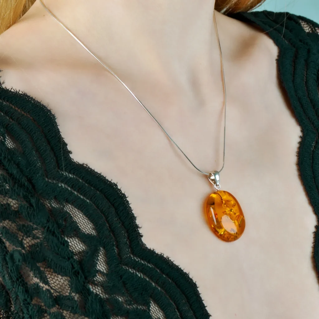 A close-up image of a genuine amber pendant showcasing its warm, golden hues. The amber is transparent with natural inclusions, capturing its organic beauty. The pendant is encased in a silver or gold frame, adding elegance to the raw, fossilized resin. The smooth surface of the amber reflects light, highlighting its natural glow and intricate details. This timeless piece of jewelry hangs delicately from a chain, blending nature and craftsmanship.