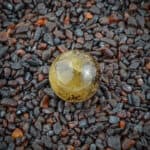 Polished yellow crystal sphere on dark gravel, showcasing natural imperfections and reflective, glossy surface.